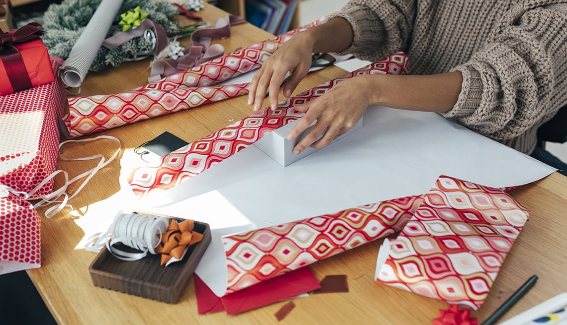 person wrapping a rectangular box with patterned red and white wrapping paper at a wooden table, surrounded by gift wrapping supplies.
