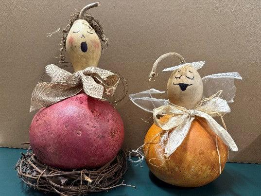 two decorative gourds painted as singing angels with closed eyes, ribbon bows, and wings, set against a plain background.