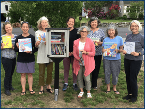 Eight women stand outdoors around a small free library, each holding children's books and smiling at the camera.