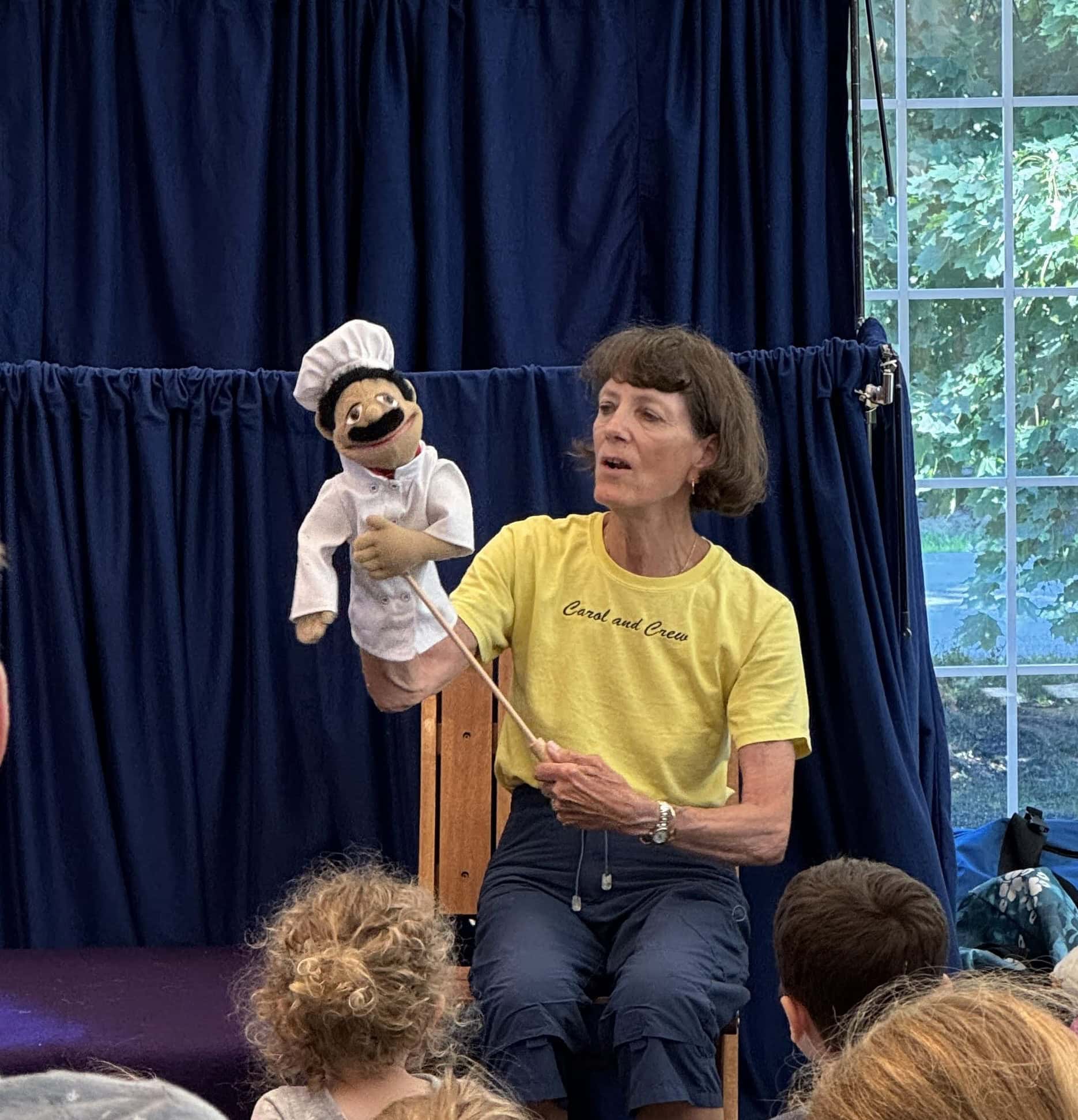 a woman in a yellow shirt performs with a chef puppet on stage in front of children, with a blue curtain and window in the background.