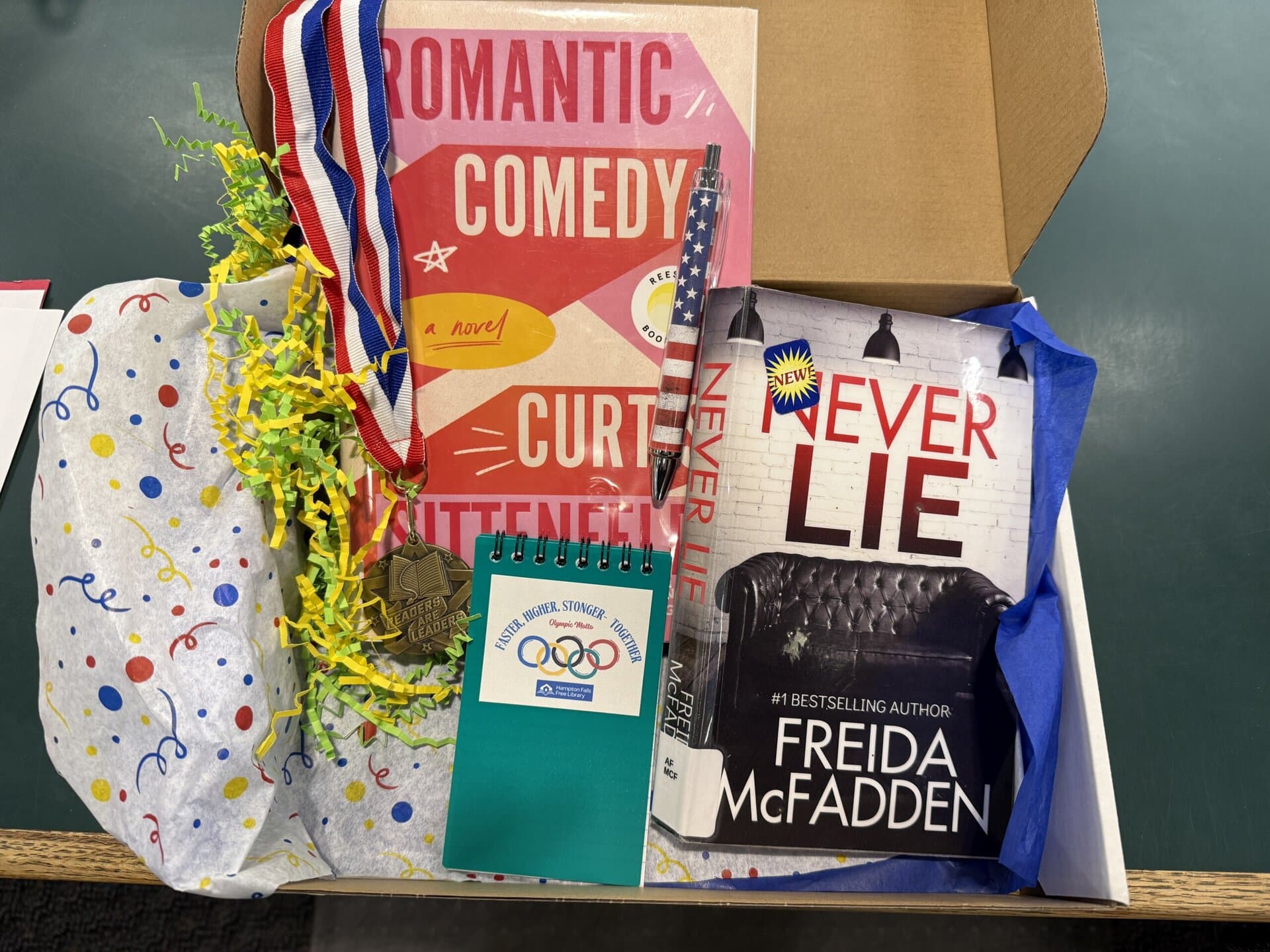 a gift box containing two books, a medal with a ribbon, a pen, colorful shredded paper, and a green notepad with an olympic themed logo.