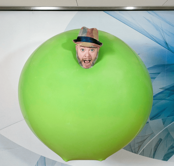 a man wearing a hat has his head poking through a large, round, green prop, with a surprised expression on his face.