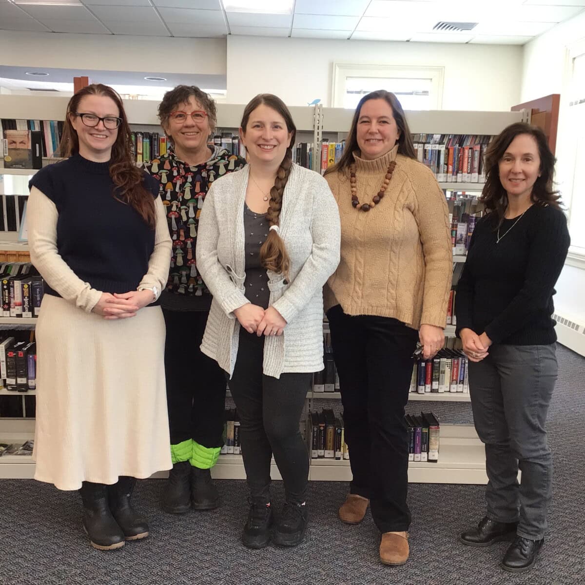 Five women standing side by side in front of bookshelves in a library, smiling at the camera.