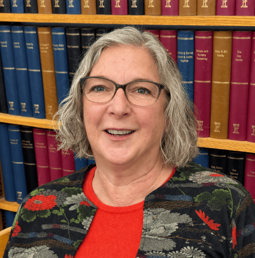 a smiling woman with gray hair and glasses wearing a floral jacket over a red shirt, standing in front of bookshelves.