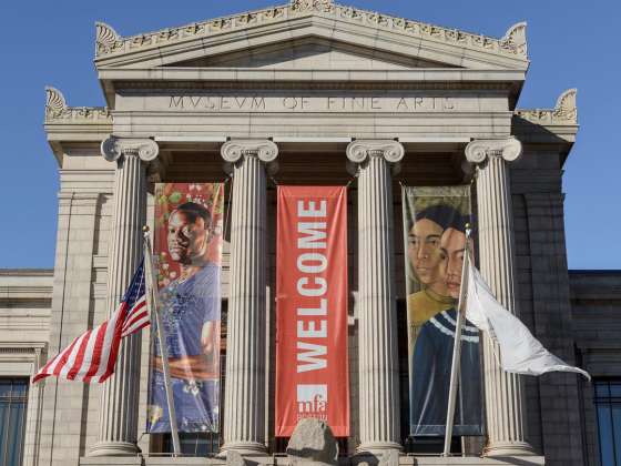 Front entrance of the Museum of Fine Arts with columns, three large banners including a red "WELCOME" banner, and American and white flags displayed outside.