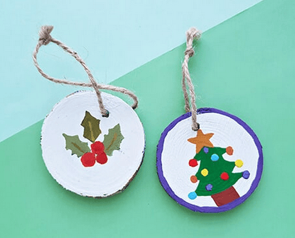 two round wooden ornaments with twine loops; one painted with holly and berries, the other with a decorated christmas tree on a green and blue background.