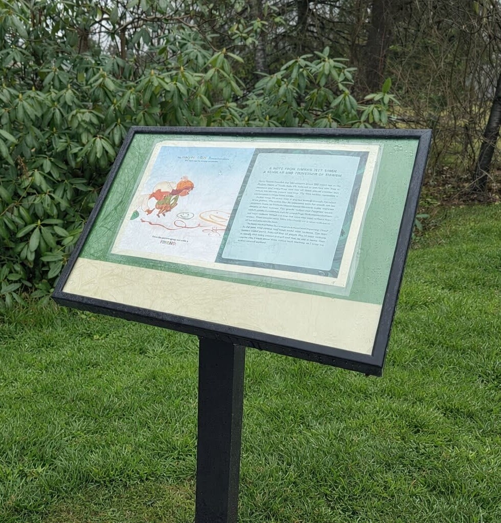 an outdoor informational sign with text and an illustration stands on a grassy area near green bushes and trees.